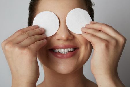 Beautiful girl model refreshing skin eyes and face with white cotton pads over gray studio background.Model with light nude make-up.Healthcare skin makeup conceptの写真素材