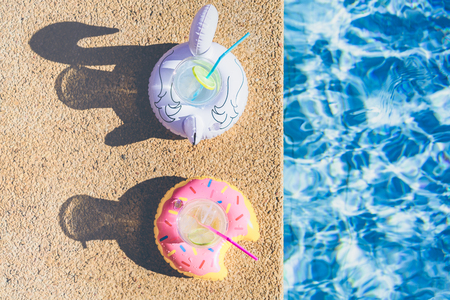 Two Tasty refreshing cocktail on inflatable donut y white flamingo plastic toy on edge of swimming pool. Ripple Water in swimming pool with sunny reflection. Top viewの写真素材