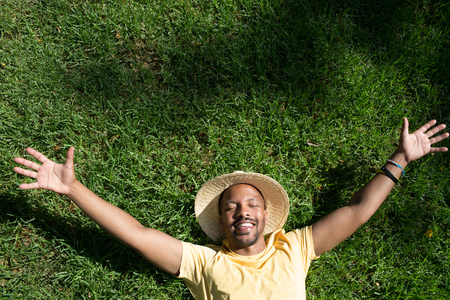 Happy African-American male model laying in the green grass with trendy straw hat and spread his hands.African man hipster treveler relaxing at park. Leisure and rest at summer dayの写真素材