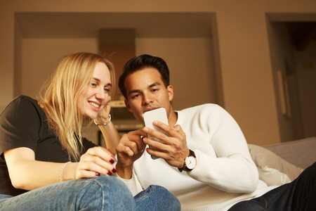 Beautiful young couple in casual clothes is using smartphones, looking at camera and smiling.の写真素材