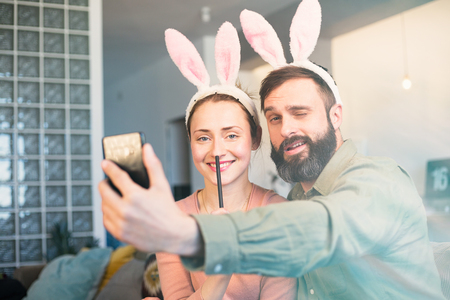 Young loving couple making selfie on mobile phone with pink rabbit ears on head. Happy family preparing for Easterの写真素材