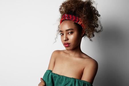 Positive attractive young female with Afro hairstyle, points aside with cheerful expression, isolated over bright light backgroud. Advertisement and ethnicity.の写真素材