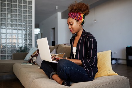 Young american african female blogger working remotely on digital netbook with internet text. African woman copywriter typing article for website on keyboard of modern laptop computer at home.の写真素材