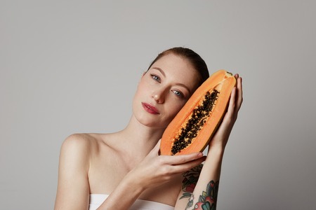 Beauty female model holding juicy papaya slice. Beautiful Joyful teen girl with freckles, funny red hairstyle and brown makeup. dvertisement and health lifestyle. Isolated on white background.の写真素材