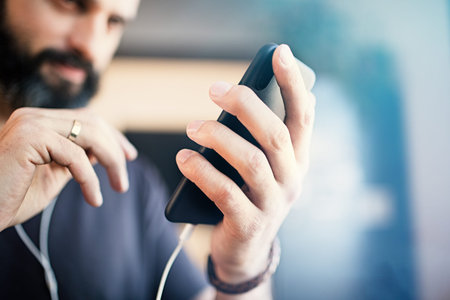 Attractive bearded guy wearing casual grey t-shirt listening music in earphones, checking social...