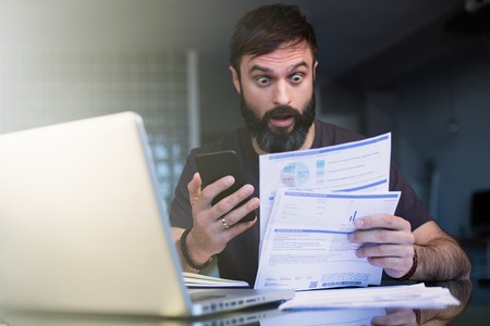 Bearded positive man working with laptop at home browsing documents . Businessman going through paperwork at home office with unpaind bills.の写真素材