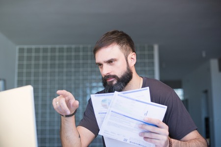 Bearded serious concentrated man working at laptop and sitting at the table while looking at the papers and holding his head with hand.の写真素材