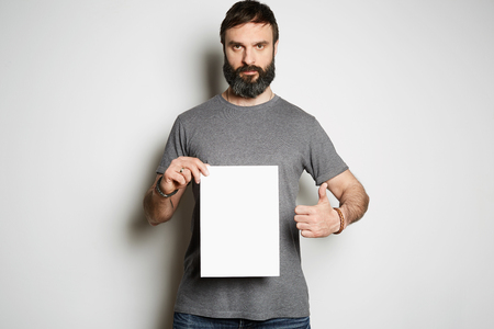 Positive brutal tattooed bearded guy holding empty sheet paper and blank gray t-shirt premium summer cotton, on white background mockup.の写真素材