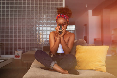 Happy young American African woman with long curly hair relaxing and drinking coffee on sofa at modern home.の写真素材