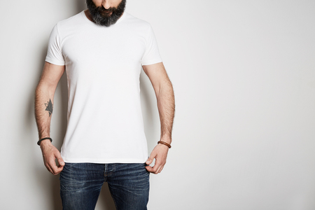 Handsome brutal tattooed bearded guy poses in blue jeans and blank gray t-shirt premium summer cotton, on white background. Mockup Copy Paste Advertisementの写真素材