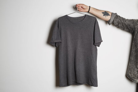 Tattooed male hand holds hang with blank gray t-shirt from premium thin cotton on white background....