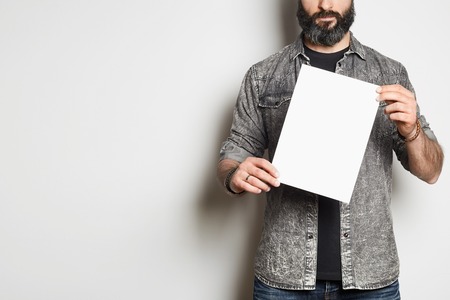 Handsome casual man in a denim shirt shows a white sheet of paper in the camera on a empty background. Mockup Copy Paste Advertisementの写真素材