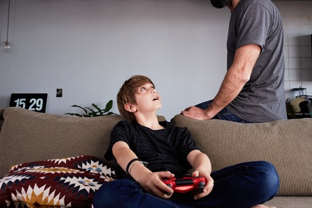 Cute young boy playing video game console seated on a sofa while his father standing close in living room at home.の写真素材