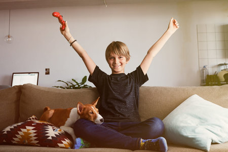 Handsome young boy celebrating his victory in video game console seated on a sofa with basenji dog...