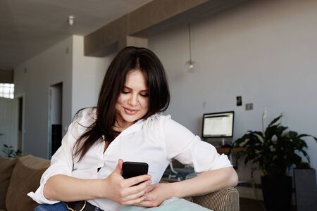 Happy Young brunette female using smartphone relaxing on sofa at living room.の写真素材