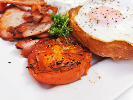 Big fried grilled tomato of English breakfast with sunny fried eggs, bacon, tomatoes, ham on Turkish flat grilled bread on white dishの写真素材