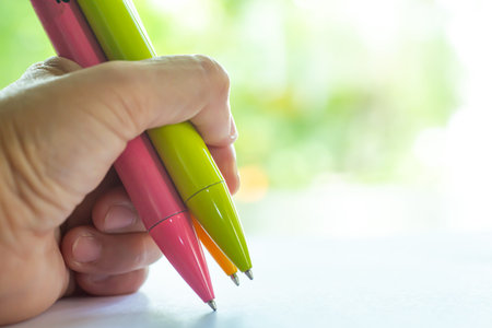 Woman's left Hand holding 3 pens ( Pink,Green, Yellow ), writing letter on white paper, Bokeh green...