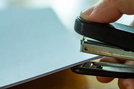 Woman's right hand holding stapler and stapling white paper on wooden background, Close up & Macro shot, Selective focus, Office Stationery conceptの写真素材
