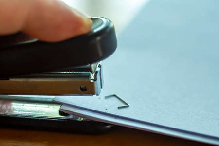 Woman's left hand holding stapler and stapling white paper on wooden background, Problem Process, Close up & Macro shot, Selective focus, Office Stationery conceptの写真素材