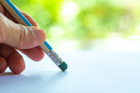 Woman's left Hand hold multicoloured pencil, Green rubber to delete from white paper, Bokeh green garden background, Notebook, Close up & Macro shot, Selective focus, Communication, Stationery conceptの写真素材