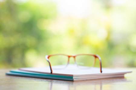 Three white notebooks with blurred orange eyeglasses on wooden table, Bokeh garden background, Close...
