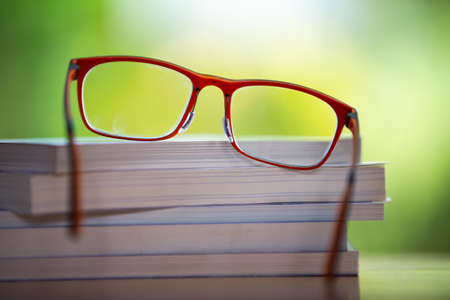 Orange eyeglasses with books on wooden table, Bokeh garden background, Close up & Macro shot, Selective focus, Stationery conceptの写真素材