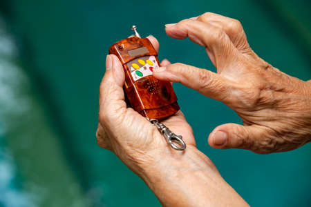 Senior woman's hands holding remote for shutter door in bokeh blue swimming pool background, Close up shot, Selective focus, Lock or Unlock, Security conceptの写真素材