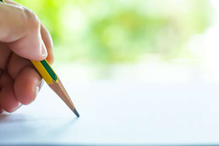 Woman's left Hand holding multicoloured pencil (Yellow & Green colour), writing letter on white paper, Bokeh green garden background, Notebook, Close up & Macro shot, Selective focus, Communication, Stationery conceptの写真素材