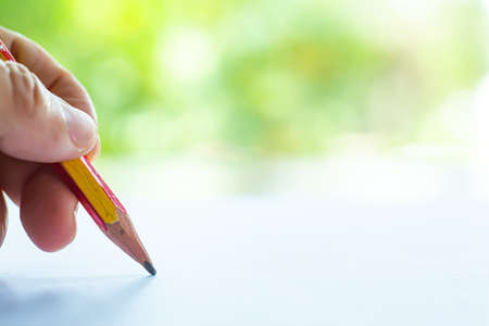 Woman's left Hand holding multicoloured pencil (Yellow & Red colour), writing letter on white paper, Bokeh green garden background, Notebook, Close up & Macro shot, Selective focus, Communication, Stationery conceptの写真素材