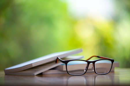 Black eyeglasses with books on wooden table, Bokeh garden background, Close up & Macro shot, Selective focus, Stationery conceptの写真素材
