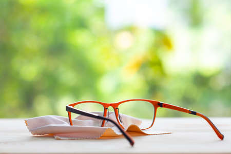 Blurred Orange & Black eyeglasses with Microfibre cleaning cloths on wooden table, In bokeh green garden background, Close up & Macro shot, Selective focus, Optical conceptの写真素材