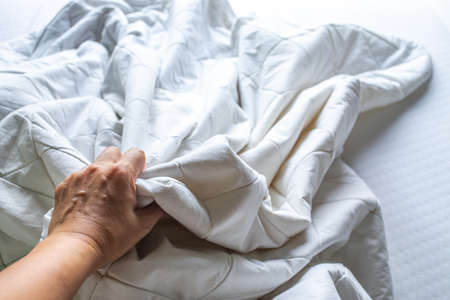 Woman's hand picking up white crumpled bed sheet on white bed, Close up shot, Selective focus, Bedroom cleaning conceptの写真素材