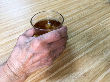 Senior woman's hand holding glass of ice Chinese tea on wooden tableの写真素材