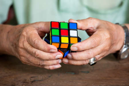 Bangkok, Thailand, 23 March 2019, Old man holding Rubik's cube and playing with it, on wooden texture background, Rubik's cube invented by a Hungarian architect Erno Rubik in 1974, Close up & Macro shot, Selective focusのeditorial素材