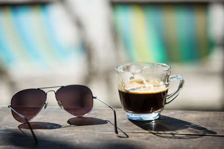 Coffee cup and Sunglasses on wood table with beach chairs & sand Backgroundの写真素材