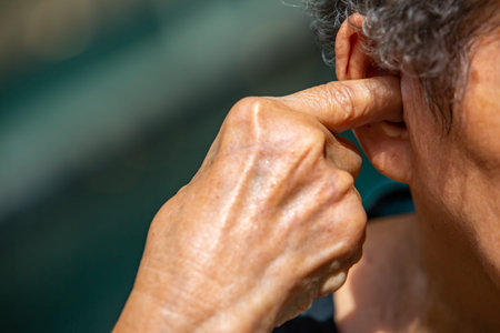 Senior woman plugging index finger into her right ear, Grey curly hairs, Swimming pool background,...