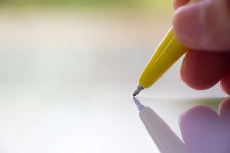 Woman's right Hand holding  yellow plastic ballpoint pen, Writing letter on white acrylic background, Close up & Macro shot, Selective focus, Communication, Business, Education, Stationery conceptの写真素材