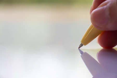 Woman's right Hand holding  beige plastic ballpoint pen, Writing letter on white acrylic background, Close up & Macro shot, Selective focus, Communication, Business, Education, Stationery conceptの写真素材