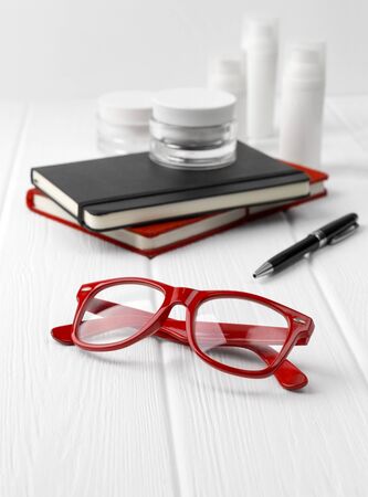 Bottles and jars of cream with black and red notepads and glasses on white wooden table. Beauty blogger conceptの写真素材