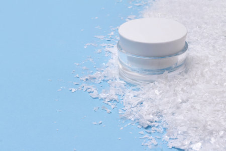 Cosmetic cream in a jar with artificial snow on blue background. Winter skin treatment concept