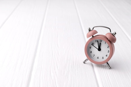Retro style pink alarm clock on white wooden background shows two minutes to twelve o'clock. Time to...