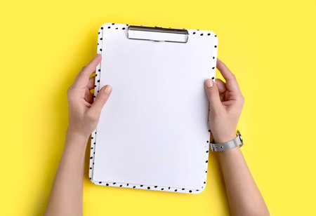 Womans hands holding modern clipboard with blank sheet on yellow background. Flat lay, top view...