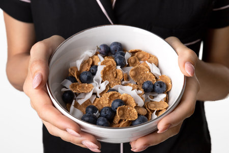 Woman in sleepwear holding bowl with whole wheat flakes, blueberry and coconut