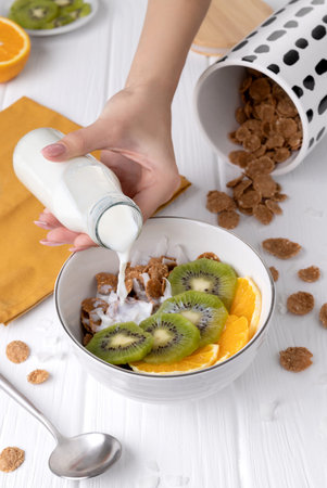 Woman pouring milk into whole wheat flakes with orange and kiwi fruits. Healthy home breakfast...