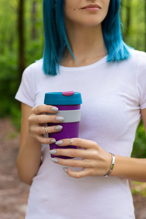 Young adult beautiful woman with blue hair standing in the outdoor garden holding a reusable cup...