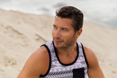 Pensive handsome young man sitting on the dunes in desert. Fashion model in trendy casual clothes...