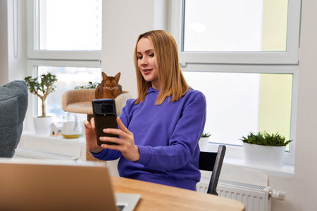 Young adult woman in violet hoodie with abyssinian cat takes a selfie. Work from home concept