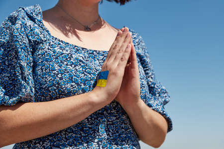 Ukrainian woman with a flag symbol on her hand prays against the blue sky. Stop warの写真素材