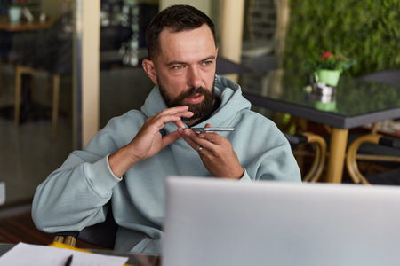 Thoughtful young adult man in casual clothes in cafe records audio messages
