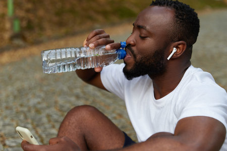 Young adult man resting on a park after a run with a bottle of water and smartphone. Outdoor workout